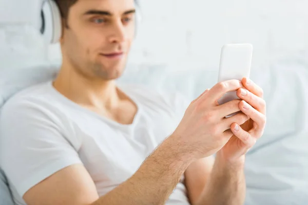 Selective focus of man in t-shirt sitting with smartphone and listening music on white background — Stock Photo