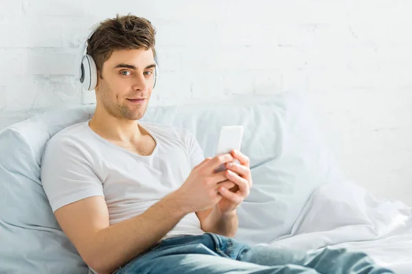 Bel homme en t-shirt blanc et jeans assis sur le lit avec smartphone et écouter de la musique dans la chambre — Photo de stock