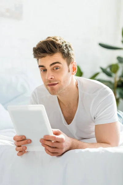 Foco seletivo de homem bonito em t-shirt branca deitada na cama com tablet digital no quarto — Fotografia de Stock
