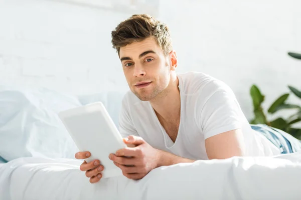 Selective focus of man in white t-shirt lying on bed with digital tablet in bedroom — Stock Photo
