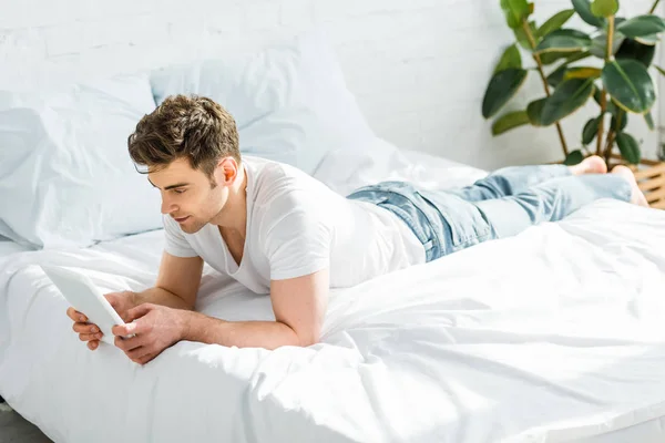 Hombre en camiseta blanca y jeans acostado en la cama con tableta digital en el dormitorio - foto de stock