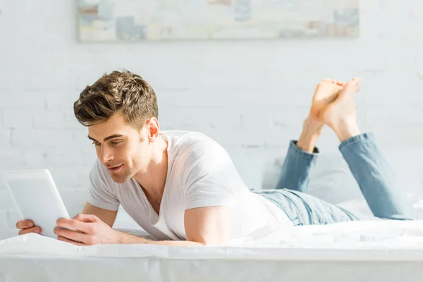 Красивый мужчина в белой футболке и джинсах на кровати с цифровым планшетом в спальне — стоковое фото