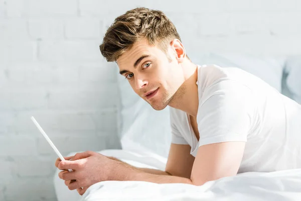 Homem bonito em t-shirt branca na cama segurando tablet digital no quarto — Fotografia de Stock