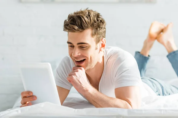 Красивий чоловік у білій футболці на ліжку тримає цифровий планшет і посміхається в спальні — стокове фото
