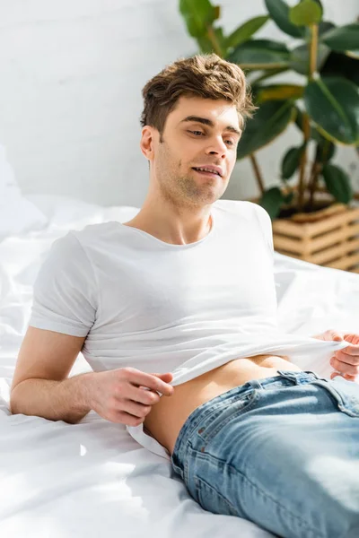 Вибірковий фокус красивого чоловіка в білій футболці і джинсах лежить на ліжку в спальні — стокове фото