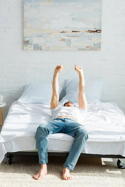 Homem de t-shirt e jeans deitado com as mãos levantadas na cama no quarto — Fotografia de Stock