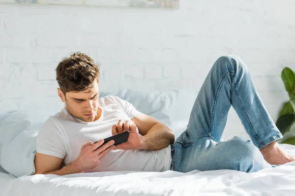 Красивый мужчина лежит на кровати и использует черный смартфон дома — стоковое фото