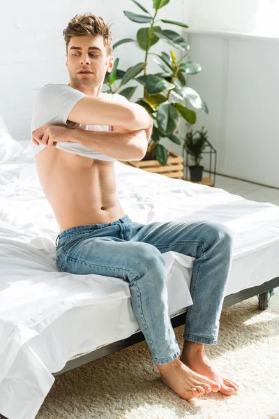 Мужчина в синих джинсах сидит на кровати и снимает футболку в спальне — стоковое фото