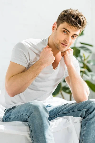 Selektiver Fokus eines gutaussehenden Mannes in weißem T-Shirt und Jeans, der auf einem Bett in der Nähe der Pflanze im Schlafzimmer sitzt — Stockfoto