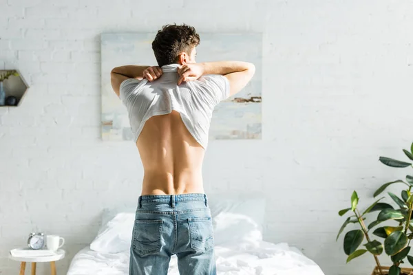 Man in blue jeans standing backwards near bed and taking off white t-shirt in bedroom — Stock Photo