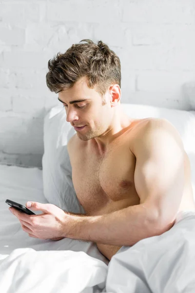 Bel homme avec torse nu tenant smartphone sur le lit à la maison — Photo de stock