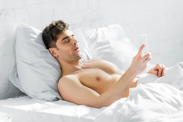 Красивый мужчина с голым туловищем лежит на кровати со смартфоном дома — стоковое фото