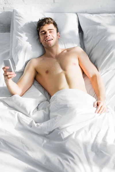 Hombre guapo con torso desnudo acostado en la cama con teléfono inteligente y sonriendo en el dormitorio - foto de stock