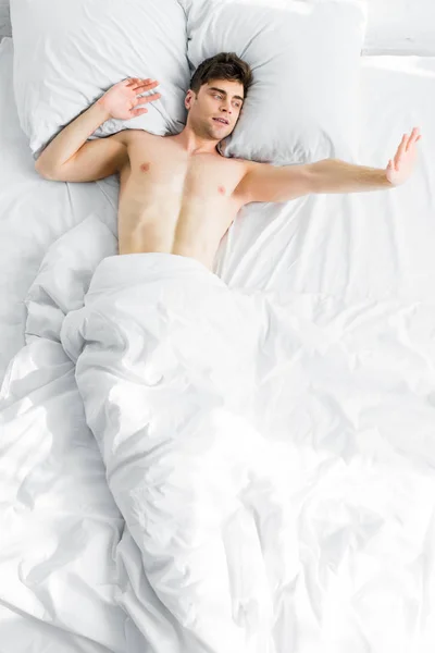 Top view of handsome man with bare torso under blanket stretching in bad at home — Stock Photo
