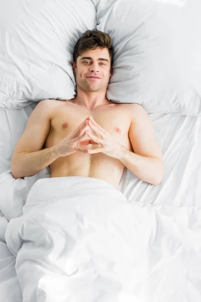Hombre guapo con el torso desnudo acostado debajo de la manta y sonriendo en la cama - foto de stock