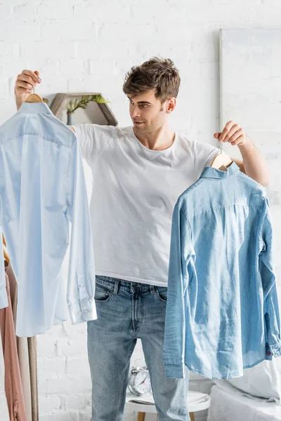 Hombre guapo en camiseta blanca sosteniendo camisas en el dormitorio - foto de stock
