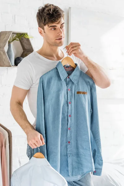 Handsome man in white t-shirt holding and trying on shirts in bedroom — Stock Photo