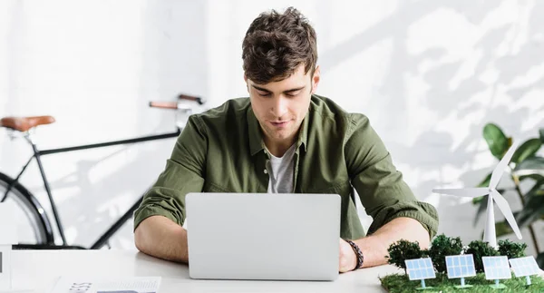 Arquitecto guapo en camisa verde sentado en la mesa con el ordenador portátil en la oficina - foto de stock