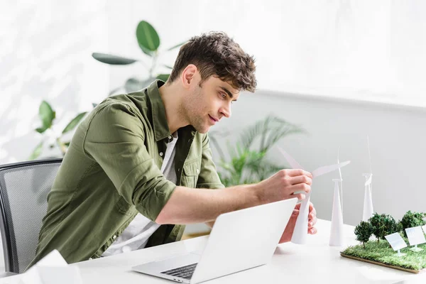 Arquiteto bonito sentado à mesa com laptop e colocando modelo de moinho de vento no escritório — Fotografia de Stock