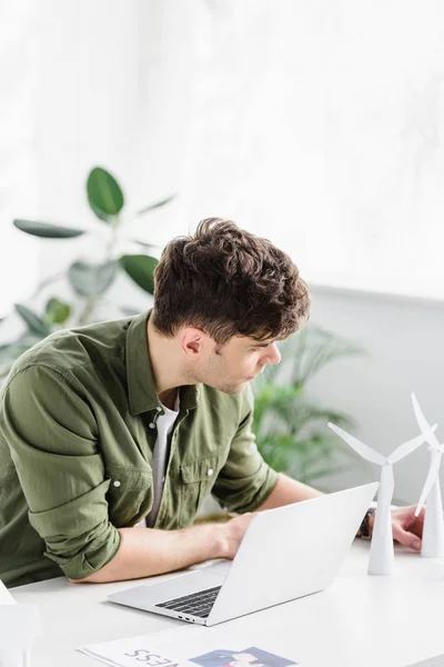 Arquiteto bonito sentado à mesa com laptop e colocando modelos de moinho de vento no escritório — Fotografia de Stock