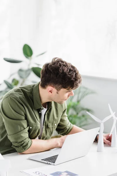 Arquitecto guapo sentado en la mesa con el ordenador portátil y poner modelos de molino de viento en la oficina - foto de stock