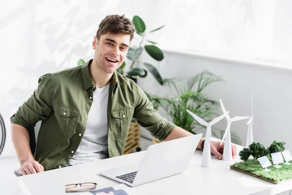 Arquiteto bonito sentado à mesa com laptop, moinhos de vento, modelos de painéis solares na grama e sorrindo no escritório — Fotografia de Stock