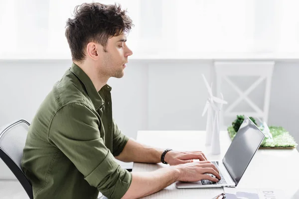 Handsome architect sitting at table and typing on laptop near windmills models in office — Stock Photo