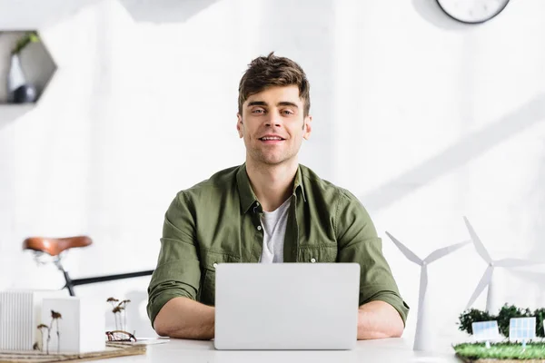 Arquitecto guapo sentado en la mesa, escribiendo en el ordenador portátil y sonriendo cerca de molinos de viento y modelos de edificios en la oficina - foto de stock