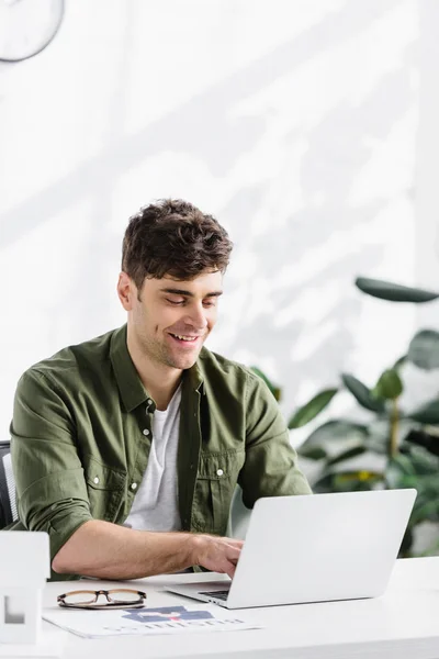 Arquitecto guapo sentado en la mesa, escribiendo en el ordenador portátil y sonriendo en la oficina - foto de stock