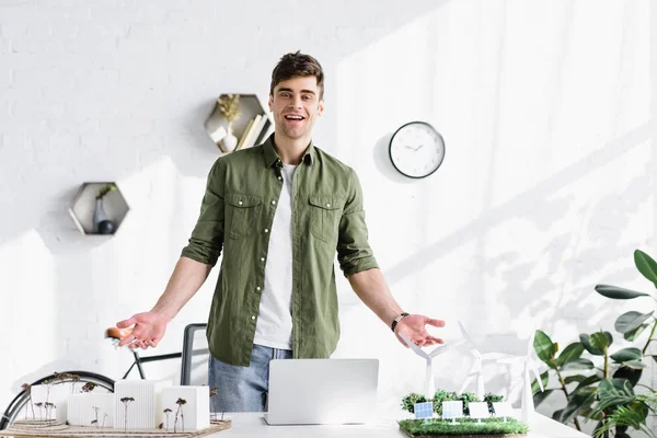 Arquitecto guapo de pie y sonriendo cerca de la mesa con árboles, edificios y molinos de viento modelos en la hierba en la oficina - foto de stock