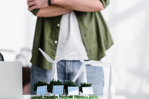 Обрезанный вид архитектора, стоящего рядом со столом с деревьями, ветряными мельницами и солнечными панелями модели на траве в офисе — стоковое фото