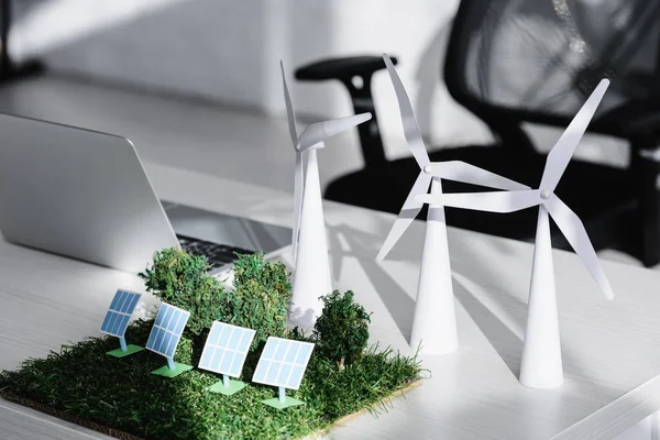 Stuhl in der Nähe Tisch mit Laptop, Bäume, Windräder, Sonnenkollektoren Modelle auf Gras im Büro — Stockfoto