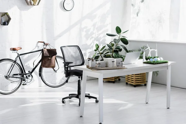 Bicicleta perto da parede de tijolo, cadeira, mesa com laptop e edifícios, árvores, modelos de moinhos de vento no escritório — Fotografia de Stock