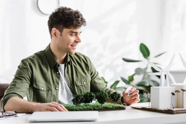 Arquiteto bonito segurando modelos de painéis solares na grama perto de laptop no escritório — Fotografia de Stock