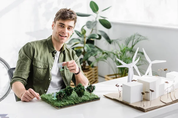 Guapo arquitecto sonriendo y sosteniendo paneles solares modelos sobre hierba en la mesa en la oficina - foto de stock