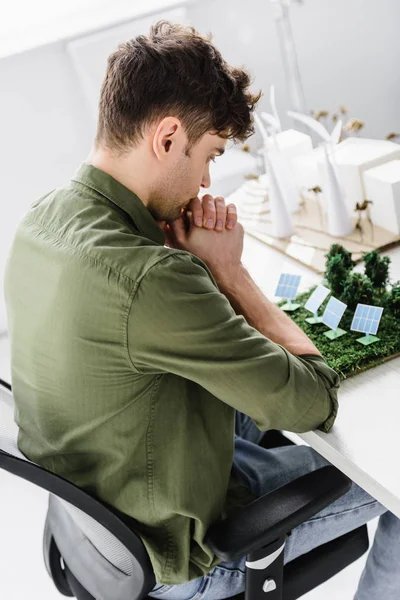 Hermoso arquitecto sentado en la silla a la mesa con árboles, molinos de viento, edificios y paneles solares modelos en la oficina — Stock Photo