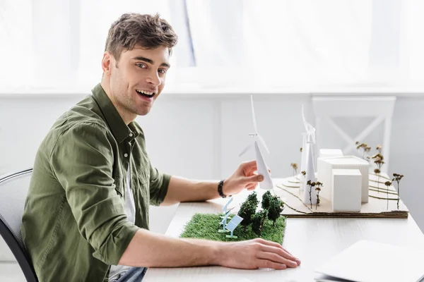 Arquitecto en camisa verde sentado a la mesa y sosteniendo el modelo de molino de viento en la oficina - foto de stock