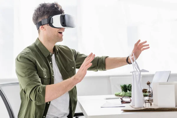 Arquiteto em realidade virtual headset sentado à mesa com moinhos de vento, edifícios e árvores modelos no escritório — Fotografia de Stock
