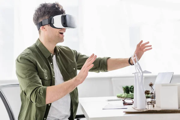 Architecte en réalité virtuelle casque assis à la table avec des moulins à vent, des bâtiments et des modèles d'arbres dans le bureau — Photo de stock