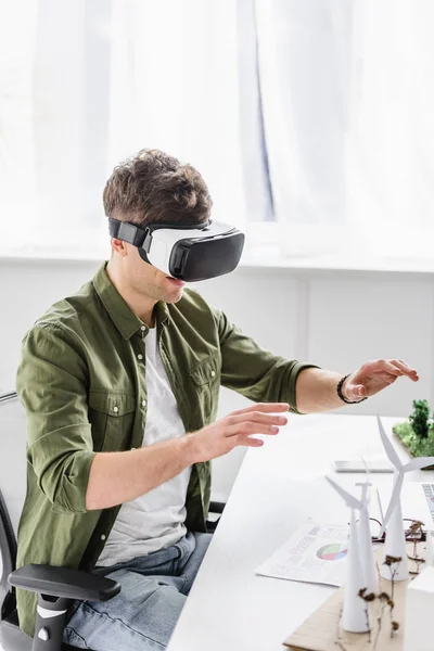 Architecte en réalité virtuelle casque assis à table avec des moulins à vent et des modèles d'arbres dans le bureau — Photo de stock