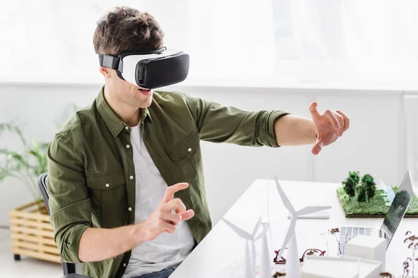 Architetto in realtà virtuale auricolare seduto a tavola con laptop, mulini a vento e alberi modelli in ufficio — Foto stock