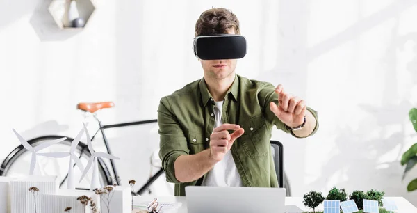 Architect in virtual reality headset at table with laptop and trees, solar panels models in office — Stock Photo