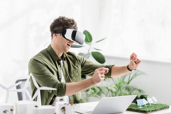 Architecte en réalité casque à table avec ordinateur portable et moulins à vent, bâtiments, panneaux solaires modèles dans le bureau — Photo de stock