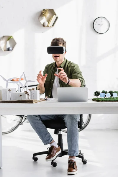 Architecte en noir casque de réalité virtuelle à table avec ordinateur portable et moulins à vent, bâtiments, arbres, panneaux solaires modèles dans le bureau — Photo de stock