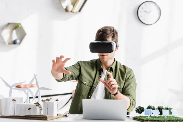 Architect in virtual reality headset sitting at table with laptop and windmills, buildings, trees, solar panels models in office — Stock Photo