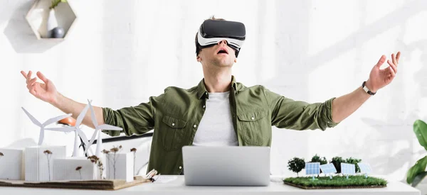 Panoramic shot of architect in reality headset at table with laptop and windmills, buildings, solar panels models in office — Stock Photo