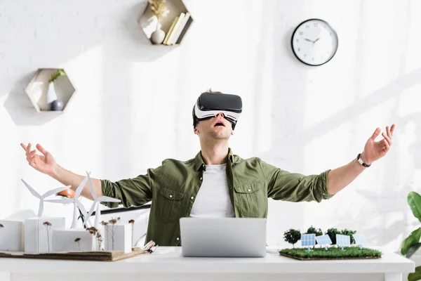 Arquitecto excitado en auriculares de realidad virtual sentado en la mesa con el ordenador portátil y los modelos en la oficina - foto de stock