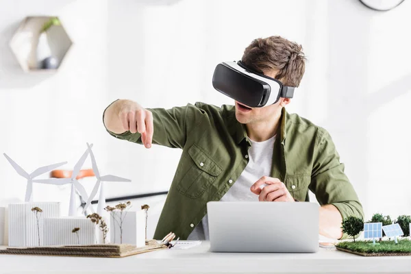 Architecte en noir casque de réalité virtuelle assis à la table avec ordinateur portable et moulins à vent, bâtiments, arbres, panneaux solaires modèles dans le bureau — Photo de stock