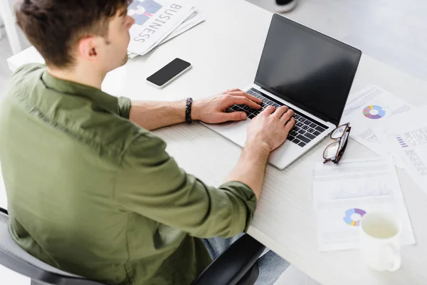 Homem de negócios bonito em camisa verde sentado à mesa com laptop e smartphone e digitando no escritório — Fotografia de Stock