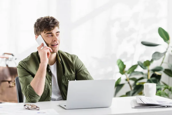 Bell'uomo d'affari in camicia verde seduto a tavola con computer portatile e parlando su smartphone in ufficio — Foto stock